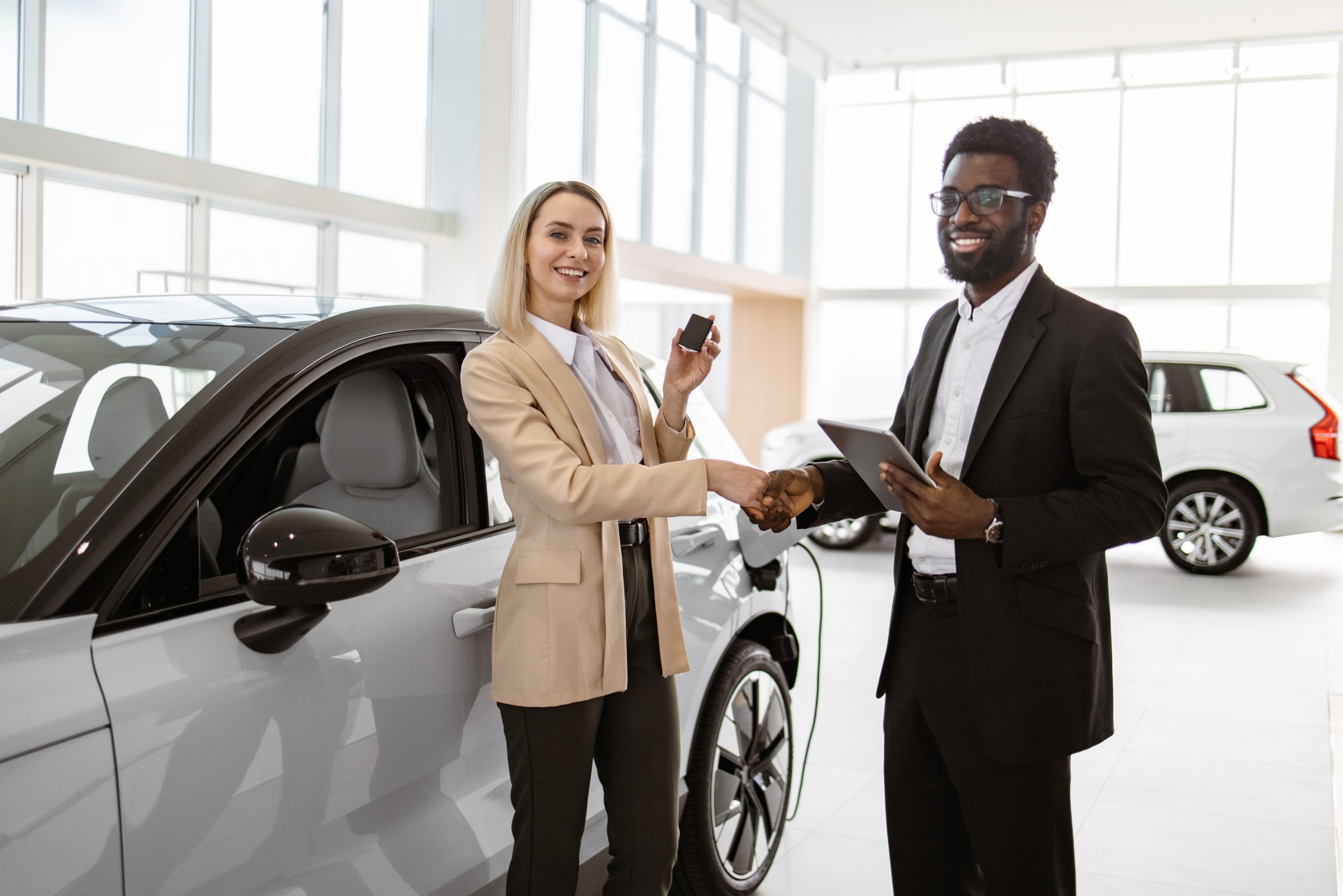 Handsome African man shaking hand with vehicle buyer, young Caucasian business lady, standing in bright dealership. Car showroom, woman purchasing new car.